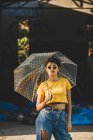 Affascinante giovane donna in abito elegante tenendo ombrello trasparente mentre in piedi sulla strada nella giornata di sole — Foto stock