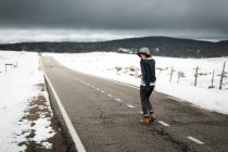 Вид на молодого человека в стильной одежде, идущего посреди асфальтовой дороги в пасмурный зимний день в красивой сельской местности — стоковое фото