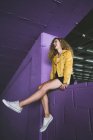 Élégante blonde bouclée en baskets et veste jaune assise sur le mur violet et riant — Photo de stock