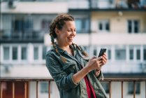 Laughing red-haired girl with braids using mobile phone against residential building — Stock Photo