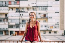 Jolie fille aux cheveux roux avec des tresses et des lunettes de soleil appuyées sur une rampe contre un immeuble résidentiel — Photo de stock