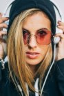 Mulher loira jovem com capuz preto e óculos de sol redondos com fones de ouvido olhando para a câmera no fundo branco — Fotografia de Stock