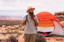 Красивий бородатий хлопець з рюкзаком, дивлячись далеко, стоячи на розмитому тлі дивовижної пустелі — стокове фото