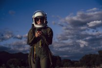 Smiling female astronaut in sunglasses holding mobile phone against evening sky — Stock Photo