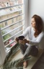 From above shot of attractive adult female in knitted sweater holding mug of hot drink and looking at street while sitting on window sill in cozy room — Stock Photo