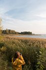 Обратный вид женщины, стоящей в высокой траве на берегу озера и фотографирующей пейзаж в осеннем солнечном свете — стоковое фото
