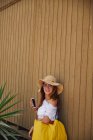Mujer sonriente en sombrero de paja con dispositivo? en la pared de madera - foto de stock