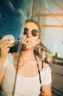 Lovely young female in stylish sunglasses blowing bubbles while sitting on city street on sunny day — Stock Photo