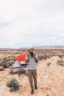Красивый бородатый мужчина с рюкзаком смотрит в сторону, когда идет рядом с палаткой в облачный день в пустыне — стоковое фото