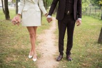 Безликий снимок элегантной модной пары, держащейся за руки и стоящей на дорожке в зеленом парке — стоковое фото
