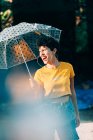 Прекрасна молода жінка в стильному вбранні тримає парасольку і дивиться геть, стоячи на вулиці під час дощу в сонячний день — стокове фото