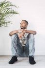 Pensive black man sitting on floor and looking away against white wall with green plant — Stock Photo