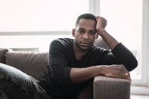 Sonhando adulto homem negro se apoiando em mãos enquanto sentado no sofá e olhando para longe na janela brilhante — Fotografia de Stock