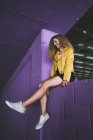 Стильний фігурні блондинка в кросівки і жовтій куртці, сидячи на purple стіни і посміхається — стокове фото