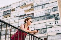 Red-haired young woman with braids using mobile phone while leaning on railing against residential building — Stock Photo
