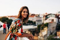 Счастливая брюнетка в красочном платье стоит на мосту и смотрит в камеру на фоне города — стоковое фото