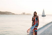 Content woman in long colorful dress walking on cobblestone promenade at sunset against seascape — Stock Photo