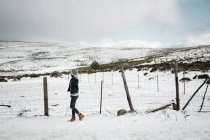 Vista laterale della giovane femmina in abiti caldi passeggiando vicino alla recinzione nella ventosa giornata invernale in una magnifica campagna — Foto stock
