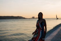 Contenu femme en robe longue et colorée marchant sur la promenade pavée au coucher du soleil contre le paysage marin — Photo de stock