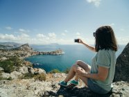 Woman taking selfie with mobile phone on cliff with seascape on background — Stock Photo