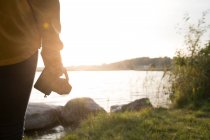 Vista posterior de la mujer de pie en la hierba alta en la orilla del lago y tomar una foto del paisaje en la luz del sol otoñal - foto de stock
