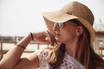 Atractiva hembra con pelo castaño en gafas de sol vestida con camisa blanca y sombrero de paja sobre fondo con muelle - foto de stock