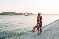 Mujer contenta en vestido largo y colorido caminando en el paseo de adoquines al atardecer contra el paisaje marino - foto de stock