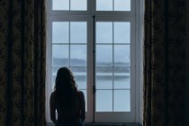 Rückansicht einer Frau, die am Fenster steht und auf den ruhigen See und die Berge unter klarem Himmel blickt — Stockfoto