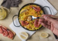 Mano humana sosteniendo tenedor sobre paella marinera tradicional española con arroz, gambas, calamares y mejillones en sartén - foto de stock