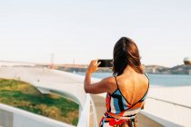 Vista posteriore della donna in abito alla moda utilizzando il telefono e scattare foto del paesaggio urbano dal ponte — Foto stock