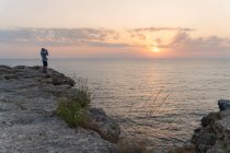 Vista lateral de um macho irreconhecível em pé em um penhasco acidentado e tirando fotos do magnífico pôr do sol sobre o mar em Tyulenovo, Bulgária — Fotografia de Stock
