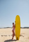 Шикарная загорелая женщина с каштановыми волосами в красном с белым купальником, держащая яркий? желтый доска для серфинга? на песчаном пляже с зелеными холмами на заднем плане — стоковое фото