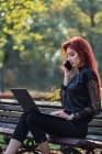 Приваблива молода жінка з ноутбуком перегляду смартфон, сидячи на лавці на сонячний день — стокове фото