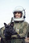 Усміхнена дівчина в старому космічному шоломі і скафандр тримає собаку в природі — стокове фото