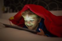 Улыбающийся мальчик смотрит мультики с цифровым планшетом в постели под одеялом — стоковое фото