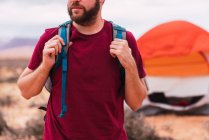 Handsome bearded guy with backpack looking away while standing on blurred background of amazing desert — Stock Photo