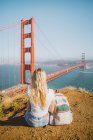 Visão traseira de uma jovem mulher sentada no chão perto da mochila e olhando para a famosa Golden Gate Bridge em São Francisco, Califórnia — Fotografia de Stock