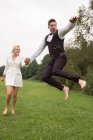 Модный взрослый жених и невеста в элегантных костюмах держатся за руки и прыгают с волнением на зеленом лугу — стоковое фото