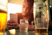 Vista da colheita do jovem afro-americano segurando e olhando para o telefone móvel, sentado no café perto de óculos com cerveja na Áustria — Fotografia de Stock
