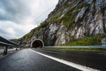 Порожня асфальтована автомагістраль, вкрита краплями дощу, що веде до тунелю в скелястому пагорбі на тлі похмурого неба — стокове фото