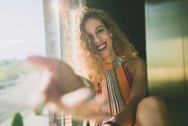 Jovem mulher encaracolado sentado em sol brilhante e estendendo a mão borrada com sorriso — Fotografia de Stock