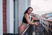 Portrait of young woman with dark hair in lingerie and black crop top standing at balcony on background of city — Stock Photo
