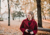Junge Dame im roten Mantel hält Digitalgerät in der Hand und blickt in die Kamera im Herbstwald — Stockfoto