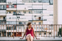 Elegante ragazza dai capelli rossi con trecce e occhiali da sole seduto vicino ringhiera contro edificio residenziale — Foto stock