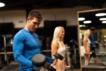 Man in blue pullover and attractive woman in sportswear standing in gym in front of mirror and doing exercise with dumbbells — Stock Photo