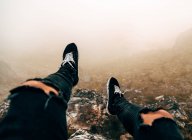 Legs of unrecognizable tourist sitting on cliff in foggy day. — Stock Photo