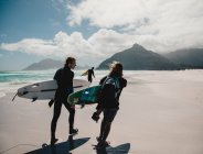 Visão traseira de pessoas com pranchas de surf andando na costa arenosa no oceano. — Fotografia de Stock