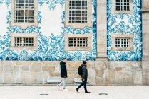 Seitenansicht fröhlicher Touristen, die an einer mit weißen und blauen Kacheln dekorierten Wand spazieren gehen. — Stockfoto