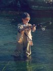 Молодий хлопчик зі стрижнем і рибальським обладнанням, що стоїть у зеленій річковій воді — стокове фото