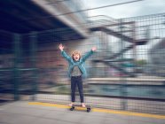 Смешной босой мальчик на скейтборде с поднятыми руками на железнодорожной платформе — стоковое фото